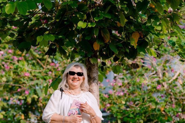Elderly woman in a white dress, smiling and enjoying the sunny day under a tree.