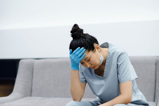 Tired nurse in scrubs sitting on a sofa, looking exhausted with her hand on her head.