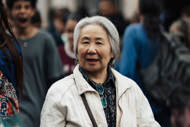 Smiling elderly woman with white hair walking in a bustling city crowd wearing a jacket.