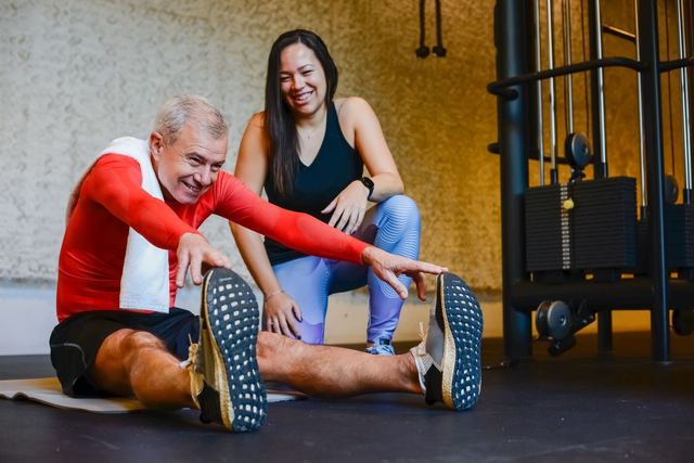 Elderly man in a gym stretching with a fitness coach, promoting a healthy lifestyle.