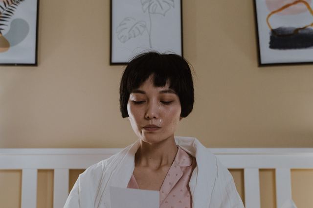 A woman in pajamas sits on a bed reading a letter, expressing emotion.