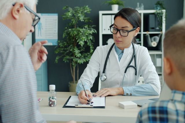 A doctor in an office consulting with an elderly man and a child, taking notes.