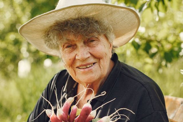 Elderly woman with a wide-brimmed hat holding fresh radishes in a sunny garden.