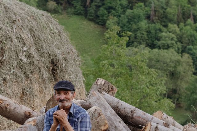 Elderly man with a dog sitting by wood logs in a lush natural setting.