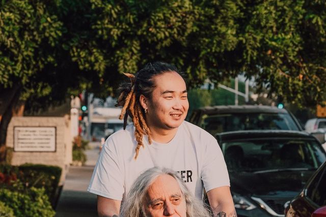 A young man pushes an elderly man in a wheelchair on a sunny sidewalk.