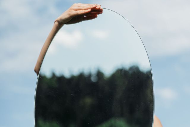 A woman in a floral dress holds an oval mirror reflecting a forest on a sunny day.