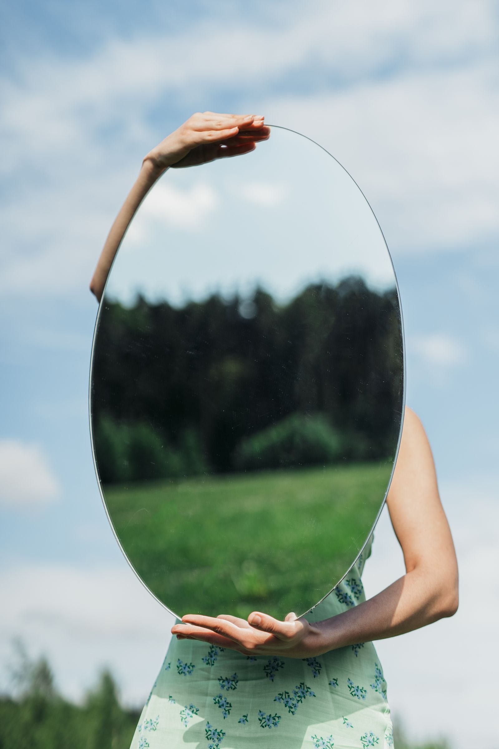 A woman in a floral dress holds an oval mirror reflecting a forest on a sunny day.